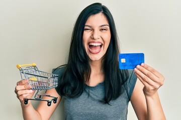 Young hispanic woman holding small supermarket shopping cart and credit card celebrating crazy and...