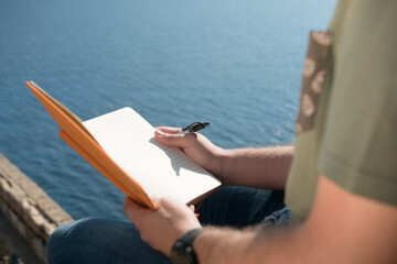 Close up of a hand taking notes on an orange noteboook in the nature in front of the sea.
