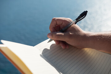 Close up of a hand taking notes on an orange noteboook in the nature in front of the sea.