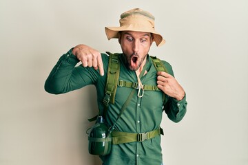 Handsome man with beard wearing explorer hat and backpack pointing down with fingers showing advertisement, surprised face and open mouth