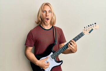 Caucasian young man with long hair playing electric guitar afraid and shocked with surprise and...