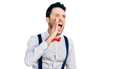 Hispanic man with beard wearing hipster look with bow tie and suspenders shouting and screaming...