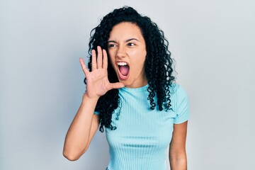 Young hispanic woman with curly hair wearing casual blue t shirt shouting and screaming loud to side with hand on mouth. communication concept.