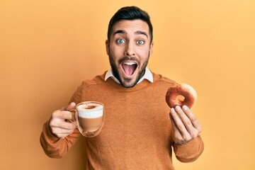 Young hispanic man eating doughnut and drinking coffee celebrating crazy and amazed for success...
