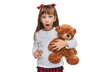 Little caucasian girl kid hugging teddy bear stuffed animal scared and amazed with open mouth for surprise, disbelief face