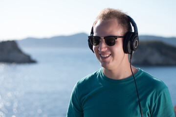 Happy young adult wearing sunglasses listening to music with headphones on a sunny day in front of the sea.