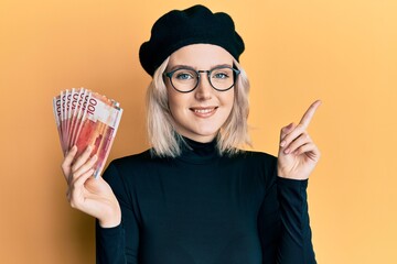 Young blonde girl holding norwegian krone banknotes smiling happy pointing with hand and finger to the side