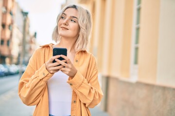 Young blonde girl smiling happy using smartphone at the city.