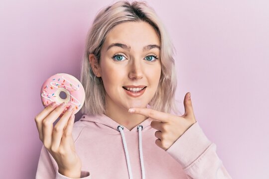 Young blonde girl holding donut smiling happy pointing with hand and finger