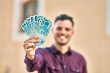 Young hispanic man smiling happy holding brazilian real banknotes at the city.