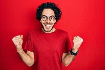 Fototapeta premium Handsome hispanic man wearing casual t shirt and glasses celebrating surprised and amazed for success with arms raised and open eyes. winner concept.