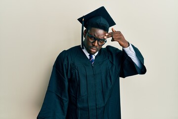 Handsome black man wearing graduation cap and ceremony robe pointing unhappy to pimple on forehead,...