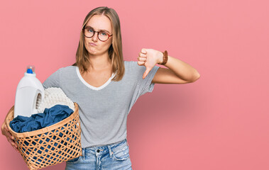 Beautiful blonde woman holding laundry basket and detergent bottle with angry face, negative sign...