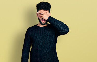 Young arab man with beard wearing casual winter sweater peeking in shock covering face and eyes with hand, looking through fingers afraid