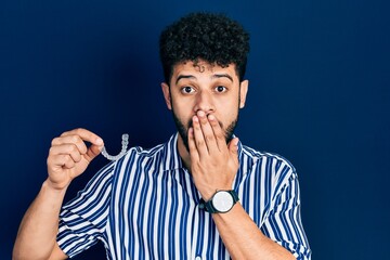 Fototapeta na wymiar Young arab man with beard holding invisible aligner orthodontic and braces covering mouth with hand, shocked and afraid for mistake. surprised expression