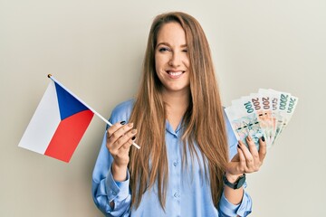 Young blonde woman holding czech republic flag and koruna banknotes winking looking at the camera...