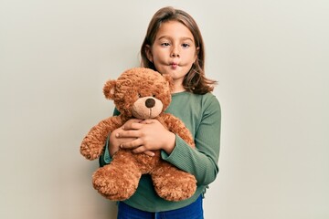 Little beautiful girl hugging teddy bear making fish face with mouth and squinting eyes, crazy and...