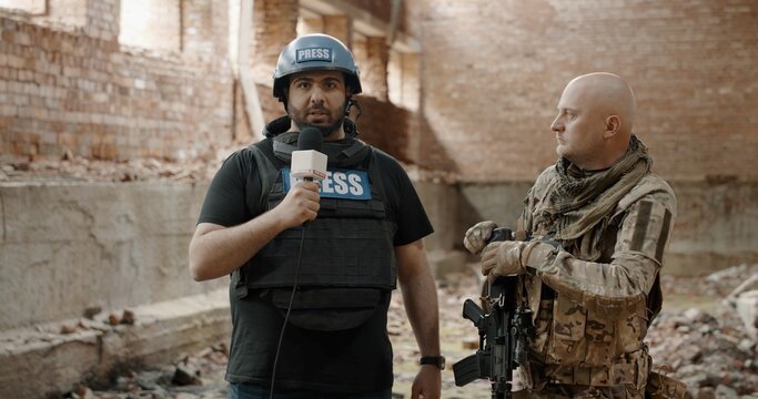 War Journalist Correspondent Wearing Bulletproof Vest And Helmet Reporting Live Near Destroyed Building, Conducting Interview With Military Representative