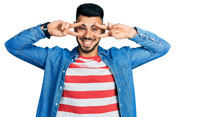 Fototapeta premium Young hispanic man with beard wearing casual denim jacket doing peace symbol with fingers over face, smiling cheerful showing victory