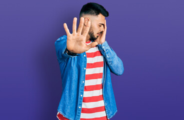 Young hispanic man with beard wearing casual denim jacket covering eyes with hands and doing stop gesture with sad and fear expression. embarrassed and negative concept.