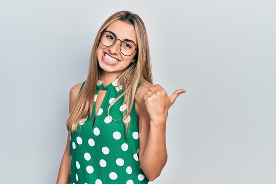 Beautiful hispanic woman wearing elegant shirt and glasses smiling with happy face looking and pointing to the side with thumb up.