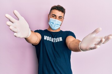 Hispanic young man wearing medical mask and volunteer t shirt looking at the camera smiling with open arms for hug. cheerful expression embracing happiness.