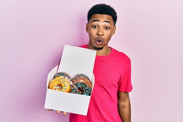 Young african american man holding box with tasty colorful doughnuts scared and amazed with open...
