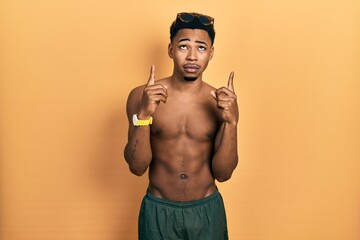 Young african american man wearing swimwear and swimmer glasses pointing up looking sad and upset, indicating direction with fingers, unhappy and depressed.