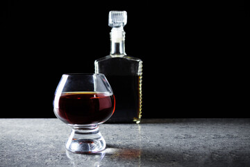 Wineglass with dark alcohol on gray granite table on black background with bottle.
