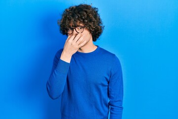 Fototapeta na wymiar Handsome young man wearing casual clothes and glasses smelling something stinky and disgusting, intolerable smell, holding breath with fingers on nose. bad smell