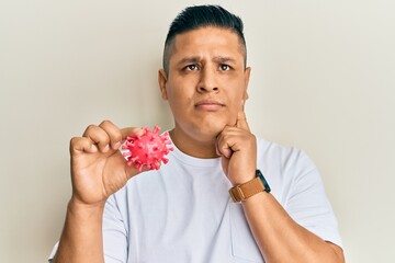 Young latin man holding virus toy serious face thinking about question with hand on chin,...