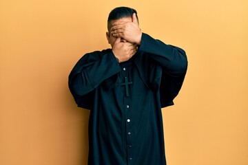 Fototapeta premium Young latin priest man standing over yellow background covering eyes and mouth with hands, surprised and shocked. hiding emotion