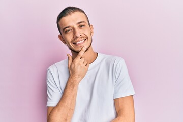 Fototapeta na wymiar Hispanic young man wearing casual white t shirt smiling looking confident at the camera with crossed arms and hand on chin. thinking positive.