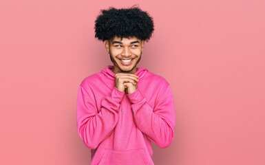 Fototapeta na wymiar Young african american man with afro hair wearing casual pink sweatshirt laughing nervous and excited with hands on chin looking to the side