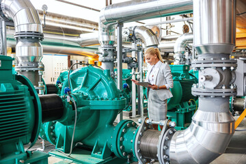 Successful smiling hardworking female manager in suit standing in heating plant with tablet in hands and checking on turbine.