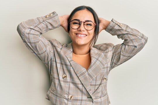 Young brunette woman wearing business jacket and glasses relaxing and stretching, arms and hands behind head and neck smiling happy