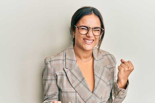 Young brunette woman wearing business jacket and glasses celebrating surprised and amazed for success with arms raised and eyes closed. winner concept.