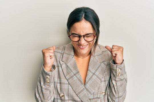 Young brunette woman wearing business jacket and glasses excited for success with arms raised and eyes closed celebrating victory smiling. winner concept.