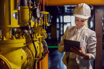 Blond dedicated successful businesswoman in formal wear checking on machinery and holding tablet...