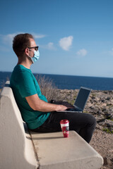 Young man wearing a face mask working on a laptop and drinking coffee sitting on a bench in a viewpoint in front of the sea. Remote working concept.