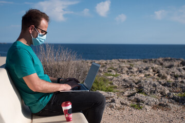 Young man wearing a face mask working on a laptop and drinking coffee sitting on a bench in a viewpoint in front of the sea. Remote working concept.