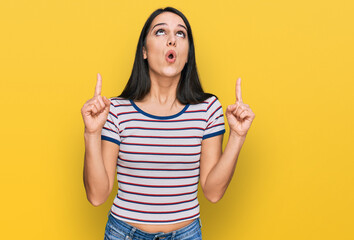 Young hispanic girl wearing casual striped t shirt amazed and surprised looking up and pointing with fingers and raised arms.