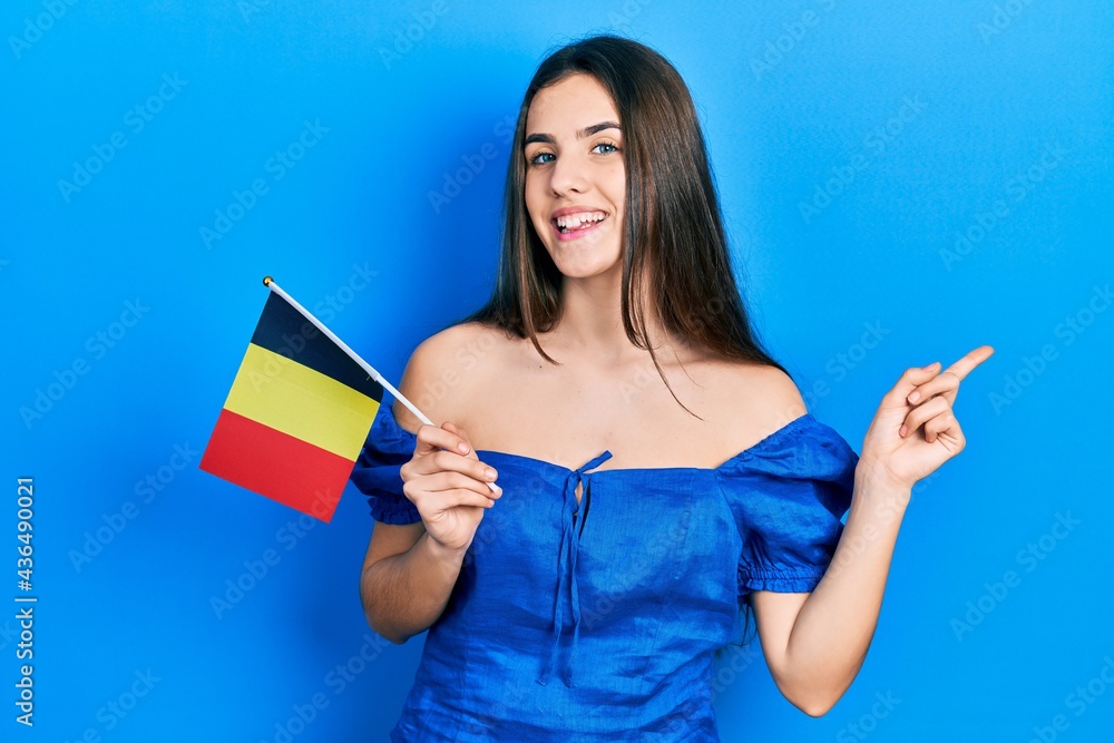 Wall mural young brunette teenager holding belgium flag smiling happy pointing with hand and finger to the side