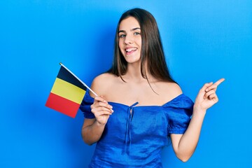Young brunette teenager holding belgium flag smiling happy pointing with hand and finger to the side