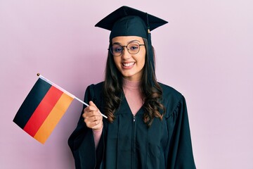 Young hispanic woman wearing graduation uniform holding germany flag looking positive and happy standing and smiling with a confident smile showing teeth