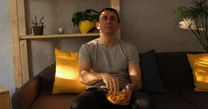 Man with a plate of crisps sits on the couch eating and watching TV. Natural evening light. Slow motion
