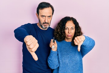 Middle age couple of hispanic woman and man holding keys of new home looking unhappy and angry...