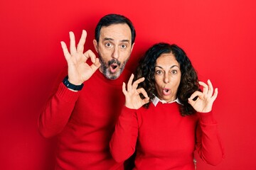 Middle age couple of hispanic woman and man hugging and standing together looking surprised and shocked doing ok approval symbol with fingers. crazy expression