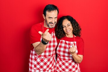 Middle age couple of hispanic woman and man wearing cook apron beckoning come here gesture with hand inviting welcoming happy and smiling