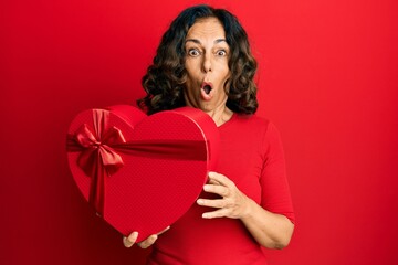 Middle age hispanic woman holding valentine day gift scared and amazed with open mouth for...